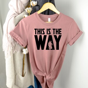 Op de afbeelding: Een stoffig roze T-shirt met de tekst "This is the Way" in zwarte letters. De tekst is in een vet, bloklettertype. Een silhouet van een persoon bevindt zich in de "Y" van het woord "Way".
