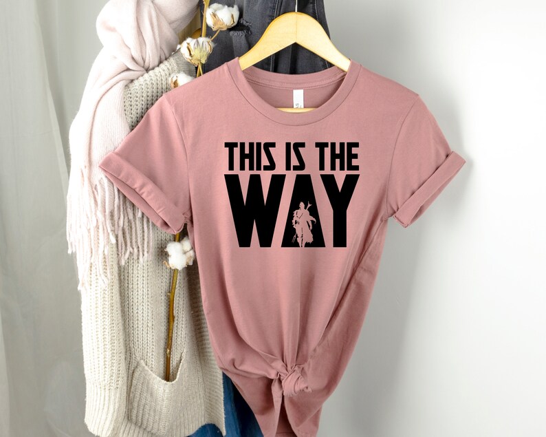 Op de afbeelding: Een stoffig roze T-shirt met de tekst "This is the Way" in zwarte letters. De tekst is in een vet, bloklettertype. Een silhouet van een persoon bevindt zich in de "Y" van het woord "Way".