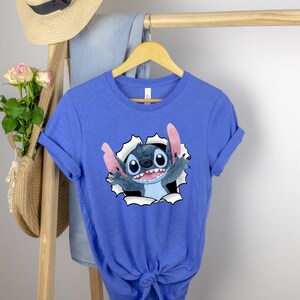 K&ouml;nnte beinhalten: Ein k&ouml;nigsblaues T-Shirt mit einer Zeichentrickfigur Stitch, die aus einem zerrissenen Loch im Stoff schaut. Stitch ist blau mit gro&szlig;en Augen und einem breiten Grinsen.