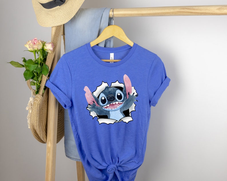 K&ouml;nnte beinhalten: Ein k&ouml;nigsblaues T-Shirt mit einer Zeichentrickfigur Stitch, die aus einem zerrissenen Loch im Stoff schaut. Stitch ist blau mit gro&szlig;en Augen und einem breiten Grinsen.