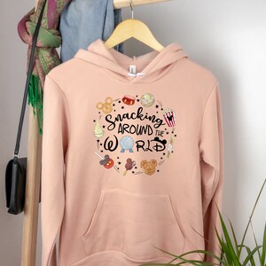 Puede incluir: Una sudadera con capucha de color melocot&oacute;n con un dise&ntilde;o gr&aacute;fico de un c&iacute;rculo con el texto "Snacking Around the World" y varios art&iacute;culos de comida y iconos con tem&aacute;tica de Disney.