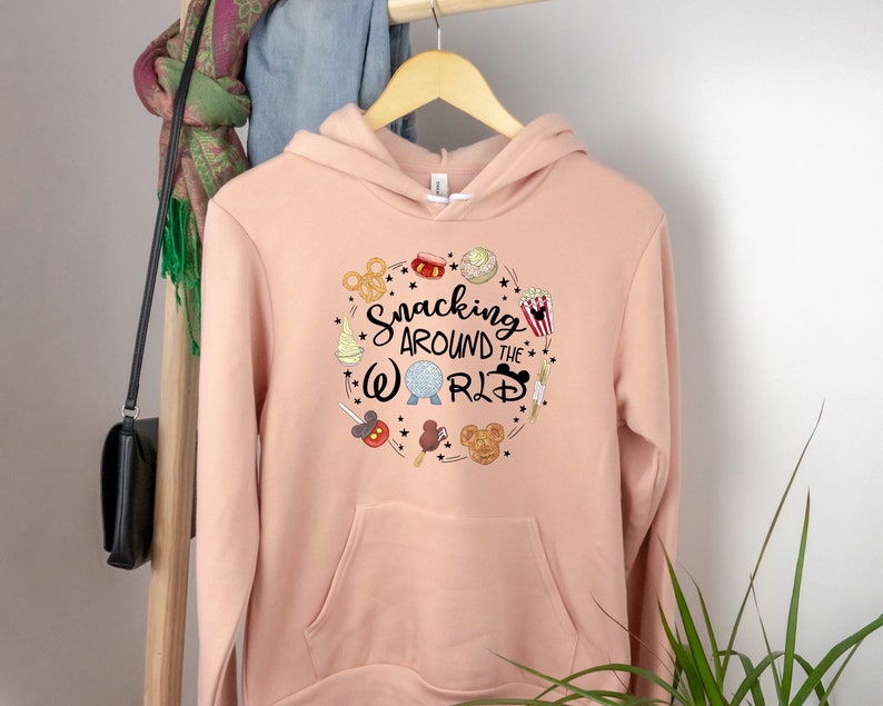 Puede incluir: Una sudadera con capucha de color melocot&oacute;n con un dise&ntilde;o gr&aacute;fico de un c&iacute;rculo con el texto "Snacking Around the World" y varios art&iacute;culos de comida y iconos con tem&aacute;tica de Disney.