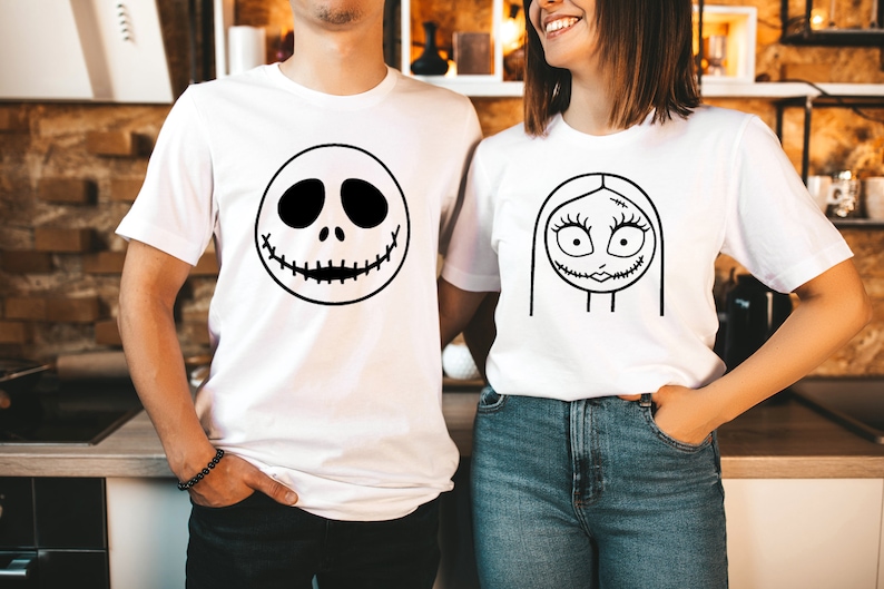 May include: A couple wearing white t-shirts with black and white cartoon faces printed on them. The man's shirt features Jack Skellington's face, while the woman's shirt features Sally's face.