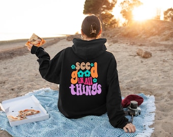 May include: A black hoodie with the text "see good in all things" in colorful, retro-style lettering. The hoodie is worn by a person sitting on a blue and white blanket on a sandy beach.