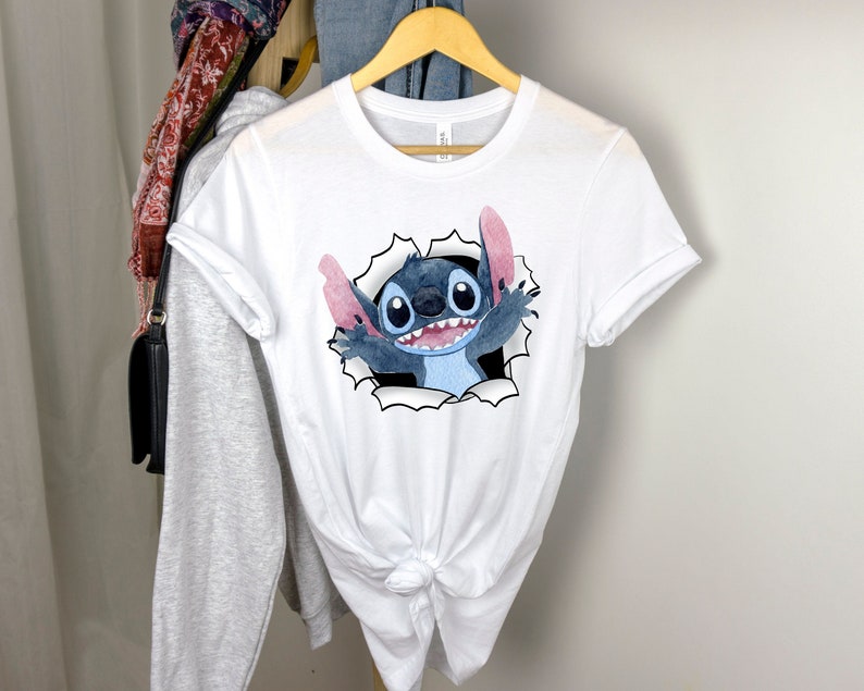 K&ouml;nnte beinhalten: Wei&szlig;es T-Shirt mit einer blauen und pinken Comicfigur von Stitch aus Lilo & Stitch, die aus einem zerrissenen Loch im Stoff schaut.