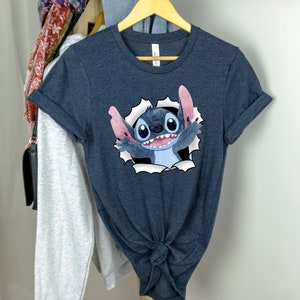 K&ouml;nnte beinhalten: Ein dunkelblaues T-Shirt mit einem Cartoon-Bild von Stitch aus Lilo & Stitch. Stitch schaut aus einem zerrissenen Loch im Shirt, mit seinem charakteristischen blauen Fell, gro&szlig;en Augen und einem breiten Grinsen.