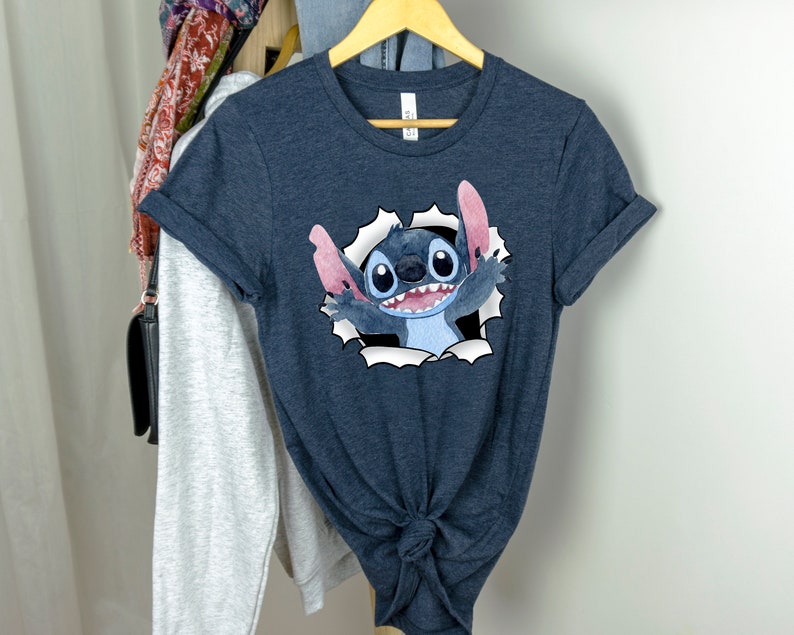 K&ouml;nnte beinhalten: Ein dunkelblaues T-Shirt mit einem Cartoon-Bild von Stitch aus Lilo & Stitch. Stitch schaut aus einem zerrissenen Loch im Shirt, mit seinem charakteristischen blauen Fell, gro&szlig;en Augen und einem breiten Grinsen.
