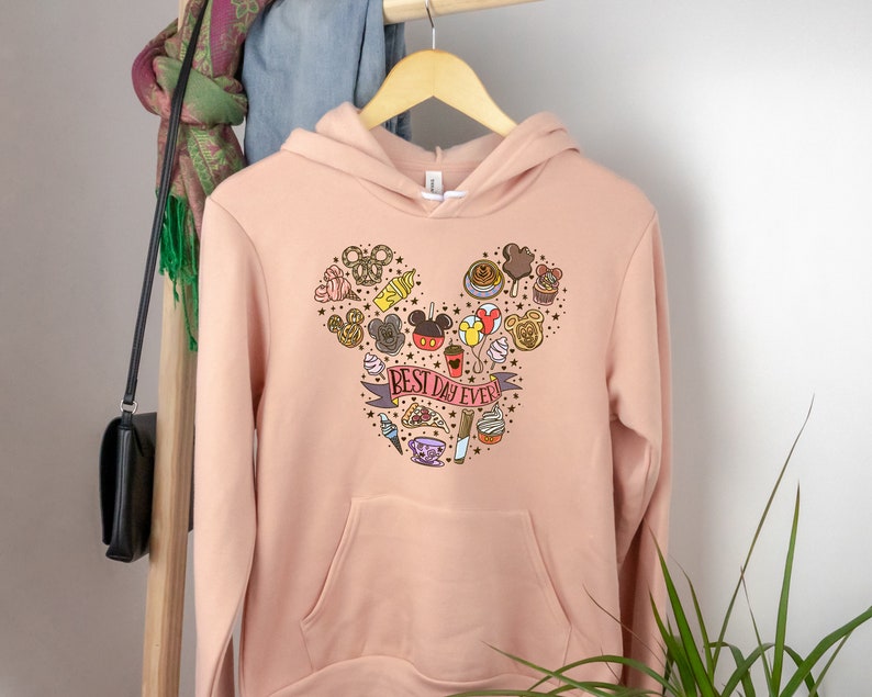 May include: A light pink hoodie with a Mickey Mouse head design featuring various Disney icons and the text "BEST DAY EVER".