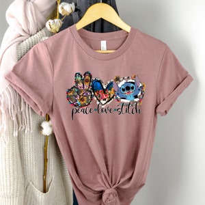 May include: A mauve t-shirt with a peace sign, heart, and Stitch from Lilo & Stitch. The peace sign and heart are surrounded by floral designs. The text "peace love stitch" is printed below the graphic.