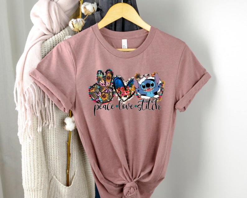 May include: A mauve t-shirt with a peace sign, heart, and Stitch from Lilo & Stitch. The peace sign and heart are surrounded by floral designs. The text "peace love stitch" is printed below the graphic.