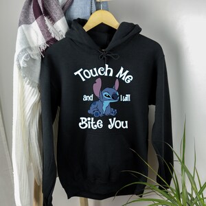 Puede incluir: Sudadera con capucha negra con un personaje azul Stitch de Lilo & Stitch. El texto en la sudadera dice "Touch Me and I Will Bite You".