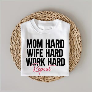 May include: White t-shirt with black text that reads "Mom Hard Wife Hard Work Hard Repeat" in a cursive font. The word "Repeat" is in pink.