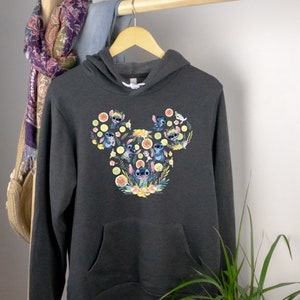 K&ouml;nnte beinhalten: Ein dunkelgrauer Kapuzenpullover mit einem Grafikdruck von Stitch-Charakteren in Form eines Mickey-Mouse-Kopfes. Der Druck zeigt Stitch-Charaktere mit Blumen und Zitrusfr&uuml;chten.