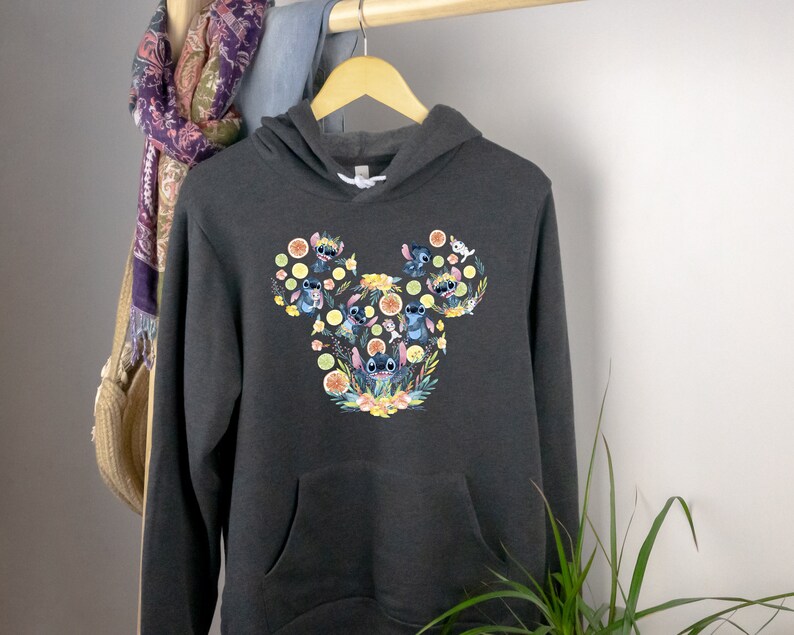 K&ouml;nnte beinhalten: Ein dunkelgrauer Kapuzenpullover mit einem Grafikdruck von Stitch-Charakteren in Form eines Mickey-Mouse-Kopfes. Der Druck zeigt Stitch-Charaktere mit Blumen und Zitrusfr&uuml;chten.