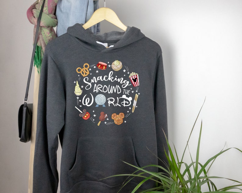Puede incluir: Una sudadera con capucha gris oscuro con un estampado gr&aacute;fico blanco de una cabeza de Mickey Mouse y el texto "Snacking Around the World" rodeado de varios bocadillos y iconos de Disney.