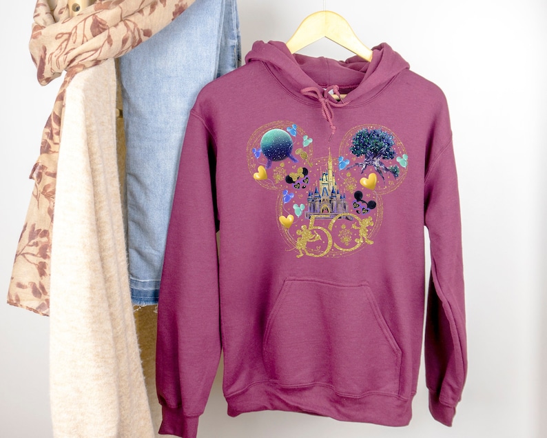 May include: Burgundy hoodie with a gold Mickey Mouse ears design featuring the Cinderella Castle, a Spaceship Earth, and a Tree of Life. The design also includes the number 50 in gold.