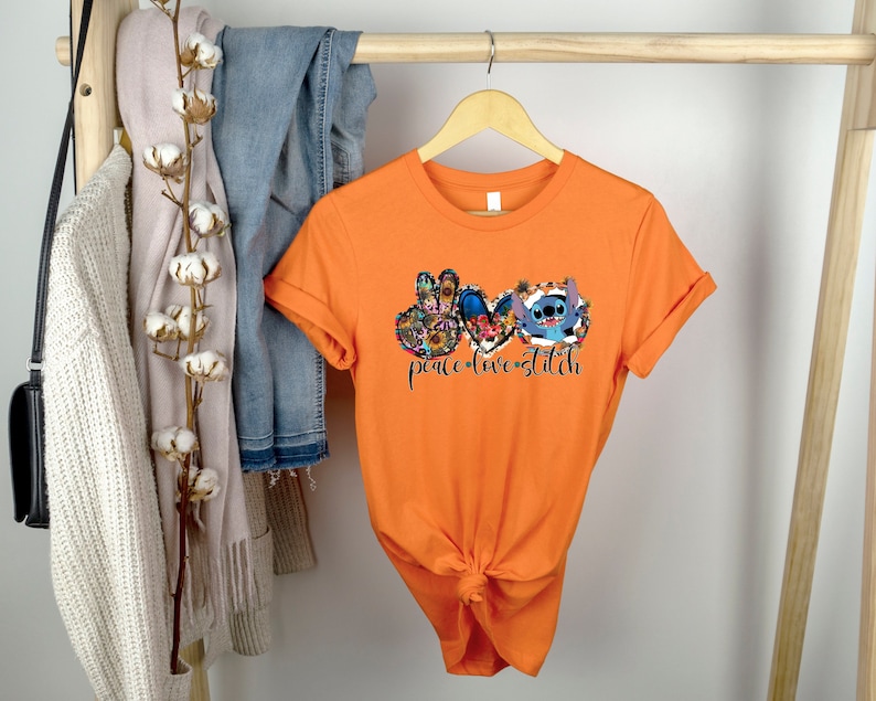 May include: Orange short-sleeved t-shirt with a graphic print of Stitch from Lilo & Stitch. The graphic features Stitch with peace signs and a heart, with the text "peace love stitch".
