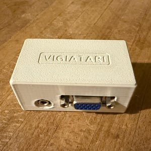 May include: A white plastic box with the text "VIGIATARI" printed on the top. The box has a VGA port, a 3.5mm audio jack, and two other ports on the side.