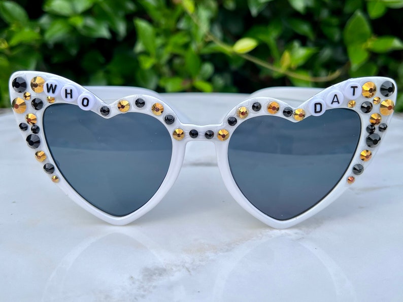May include: White heart-shaped sunglasses with black and gold rhinestones and the words "WHO DAT" on the frames.