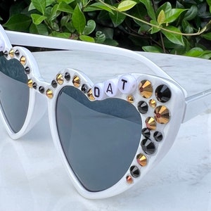 May include: White heart-shaped sunglasses with black and gold rhinestones and the words "WHO DAT" spelled out in white beads.