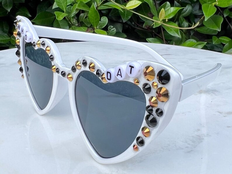May include: White heart-shaped sunglasses with black and gold rhinestones and the words "WHO DAT" spelled out in white beads.