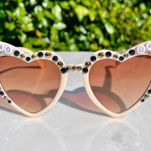 May include: Heart-shaped sunglasses with a white frame and brown lenses. The sunglasses are decorated with black and gold rhinestones and the words "WHO DAT" are spelled out in white letters on the frames.