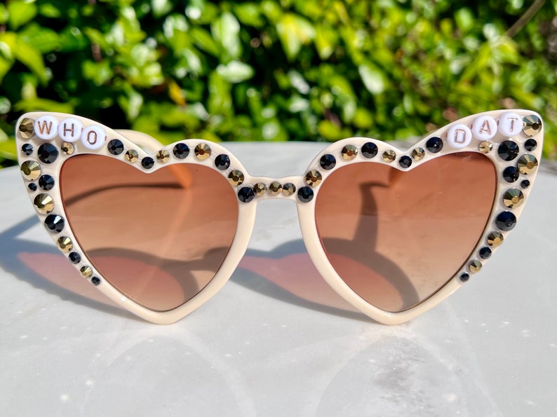 May include: Heart-shaped sunglasses with a white frame and brown lenses. The sunglasses are decorated with black and gold rhinestones and the words "WHO DAT" are spelled out in white letters on the frames.