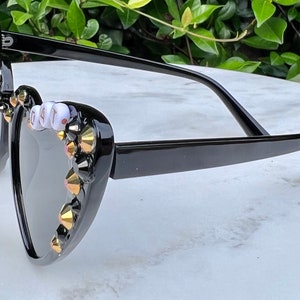 May include: Black heart-shaped sunglasses with gold and black rhinestones and white beads spelling "G.A.T."