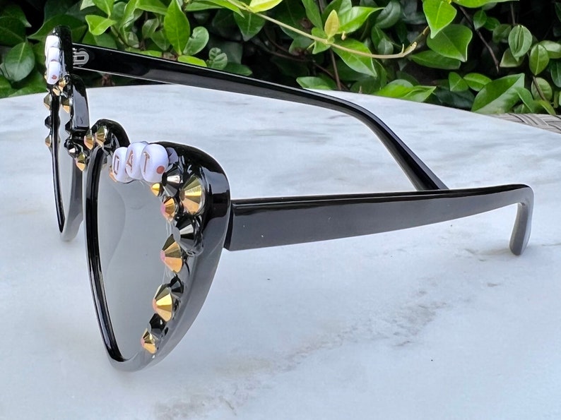 May include: Black heart-shaped sunglasses with gold and black rhinestones and white beads spelling "G.A.T."