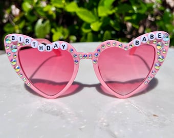 May include: Pink heart-shaped sunglasses with pink lenses. The frames are adorned with iridescent rhinestones and the words "BIRTHDAY BABE" spelled out in white letter beads. A fun and festive accessory.