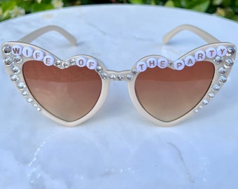 May include: White heart-shaped sunglasses with rhinestones and the words "Wife of the Party" in white letters.