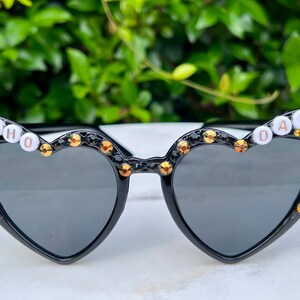 May include: Black heart-shaped sunglasses with gold rhinestones and white letters spelling "WHO DAT" on the frames.