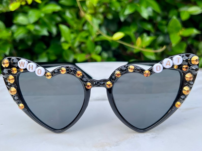 May include: Black heart-shaped sunglasses with gold rhinestones and white letters spelling "WHO DAT" on the frames.