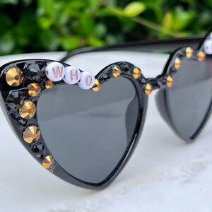 May include: Black heart-shaped sunglasses with gold and black rhinestones and the word "WHO" spelled out in white beads.