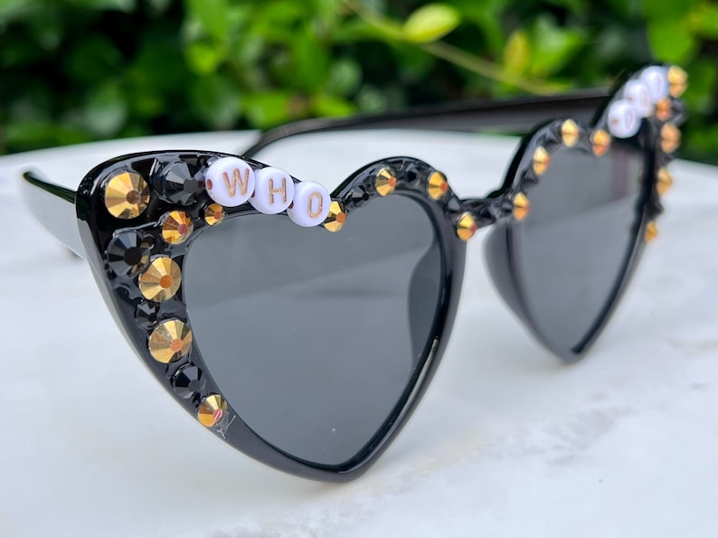 May include: Black heart-shaped sunglasses with gold and black rhinestones and the word "WHO" spelled out in white beads.