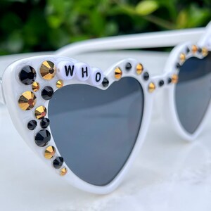 May include: White heart-shaped sunglasses with black lenses and rhinestones. The rhinestones are gold and black. The sunglasses have the word "WHO" spelled out in white letters on the frame.