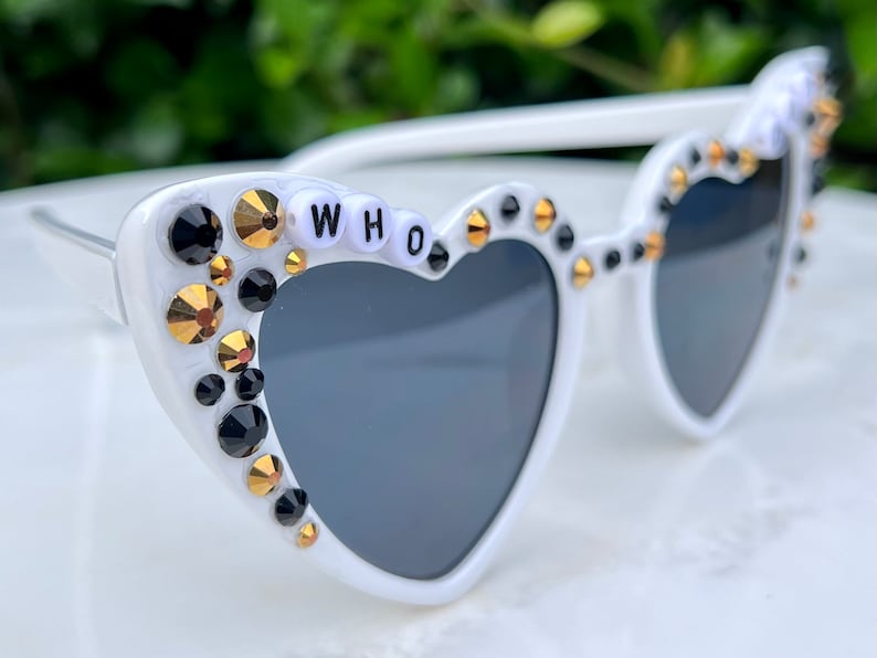 May include: White heart-shaped sunglasses with black lenses and rhinestones. The rhinestones are gold and black. The sunglasses have the word "WHO" spelled out in white letters on the frame.