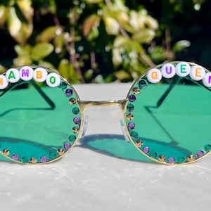 May include: Round green sunglasses with gold frames and rhinestones. The sunglasses have white beads with the words "MAMBO QUEEN" spelled out on the frames.