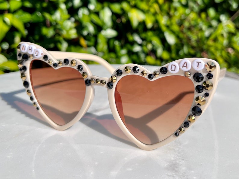 May include: White heart-shaped sunglasses with black and gold rhinestones and the words "WHO DAT" on the frames.