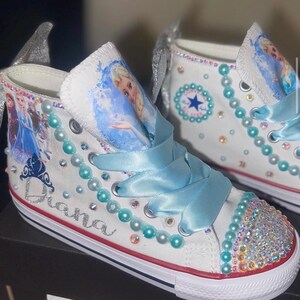 May include: A pair of white high-top sneakers featuring light blue ribbon laces, iridescent rhinestones on the toe caps, and turquoise pearl accents. The sides display character graphics, a silver glitter crown, and the name Diana in script.
