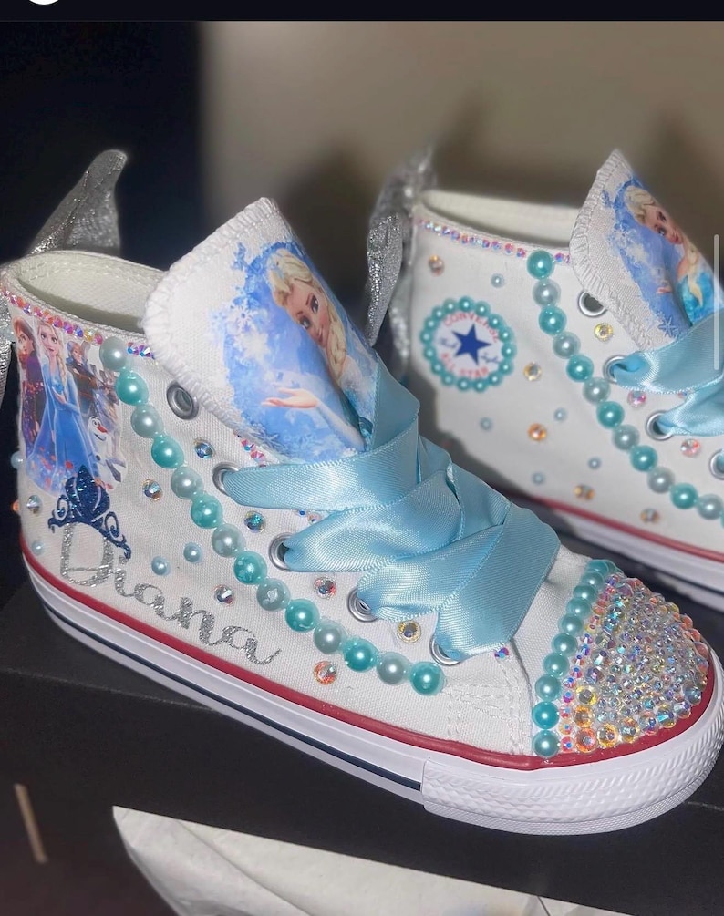May include: A pair of white high-top sneakers featuring light blue ribbon laces, iridescent rhinestones on the toe caps, and turquoise pearl accents. The sides display character graphics, a silver glitter crown, and the name Diana in script.