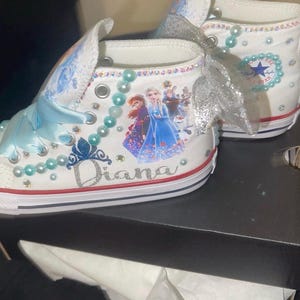 May include: White high-top sneakers featuring light blue ribbon laces, silver glitter bows, and light blue pearl accents. The side displays a graphic of characters from Frozen, a blue glitter crown, and the name Diana in silver glitter script.