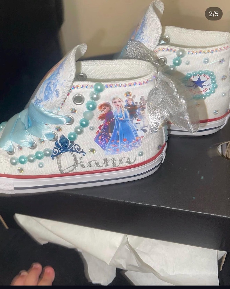 May include: White high-top sneakers featuring light blue ribbon laces, silver glitter bows, and light blue pearl accents. The side displays a graphic of characters from Frozen, a blue glitter crown, and the name Diana in silver glitter script.