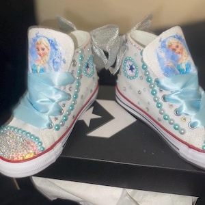 May include: A pair of white high-top sneakers featuring a character graphic of a blonde figure in a blue dress. The shoes are decorated with light blue ribbon laces, rows of light blue pearls, clear rhinestones on the toe, and a silver bow.