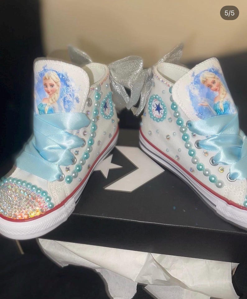 May include: A pair of white high-top sneakers featuring a character graphic of a blonde figure in a blue dress. The shoes are decorated with light blue ribbon laces, rows of light blue pearls, clear rhinestones on the toe, and a silver bow.
