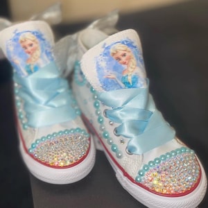 May include: A pair of white high-top sneakers featuring a graphic of Elsa from Frozen on the tongue. The shoes are decorated with light blue satin ribbon laces, rows of light blue pearls along the eyelets, and iridescent rhinestones on the toe.