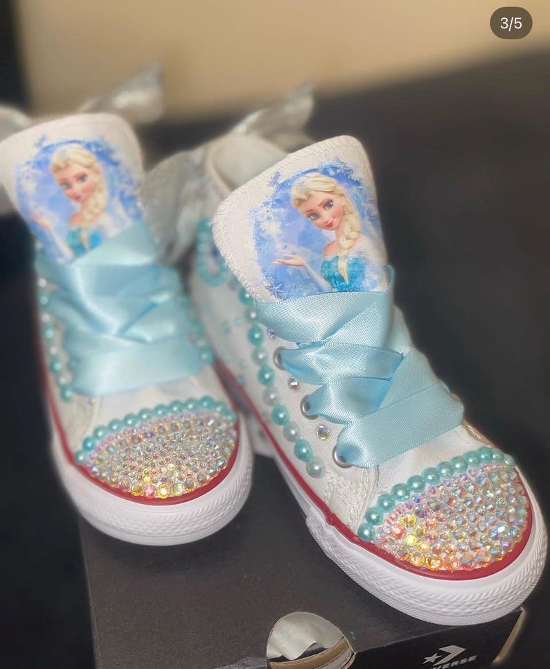 May include: A pair of white high-top sneakers featuring a graphic of Elsa from Frozen on the tongue. The shoes are decorated with light blue satin ribbon laces, rows of light blue pearls along the eyelets, and iridescent rhinestones on the toe.