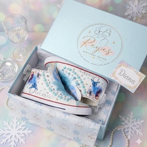 May include: A pair of white high-top sneakers decorated with blue ribbon laces, light blue pearls, and images of characters from Frozen. The shoes feature the name Diana in silver glitter and are presented in a light blue box with the Reyes logo.