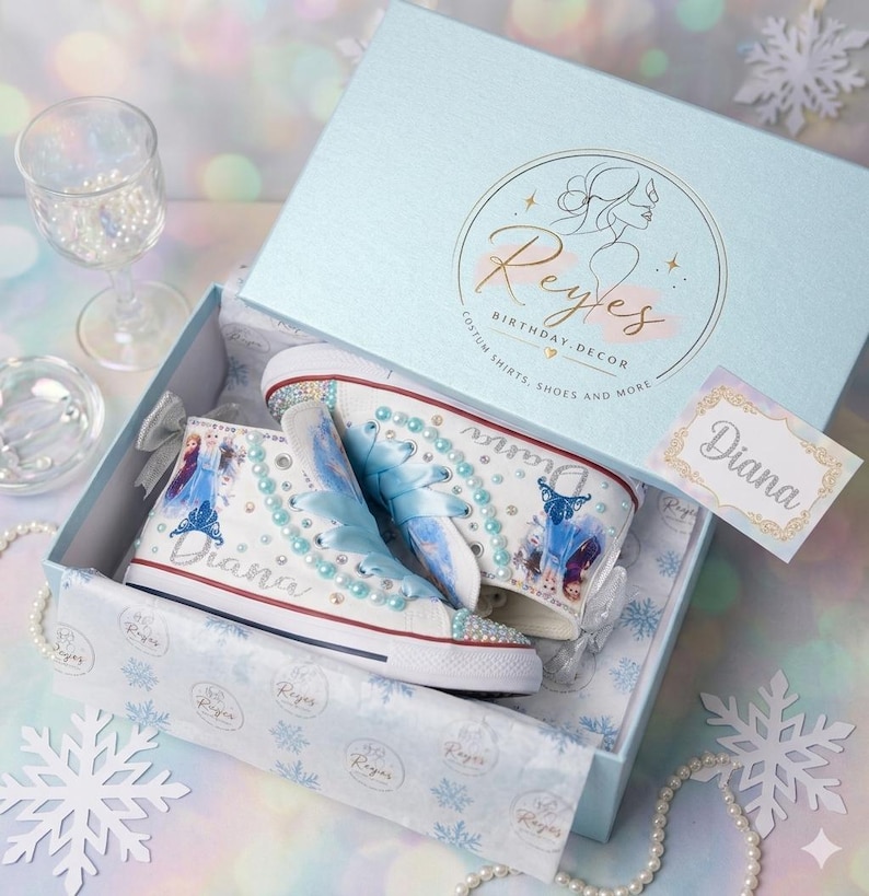 May include: A pair of white high-top sneakers decorated with blue ribbon laces, light blue pearls, and images of characters from Frozen. The shoes feature the name Diana in silver glitter and are presented in a light blue box with the Reyes logo.