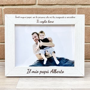 Può includere: Una cornice bianca con una foto di un padre e un figlio. La cornice ha un'iscrizione personalizzata che dice "Tanti auguri papà, sei la persona che mi ha insegnato a sorridere Ti voglio bene Il mio papà Alberto".